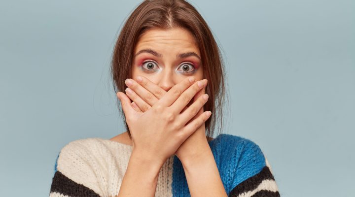 Young woman covering her mouth with hands.Scared girl puts palms on mouth,keeps silence,isolarted on blue background